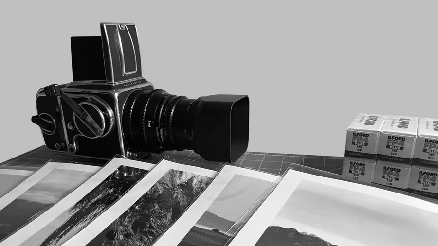 A Hasselblad camera on a cutting mat beside boxes of Ilford FP4 Plus 120 film, with four finished silver gelatin prints laid out in front of it.
