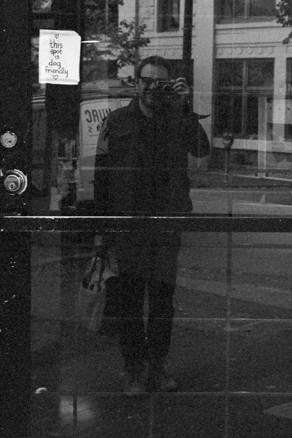 The photographer reflected in a shop window, camera raised to his eye, beside a hand-lettered sign reading “this spot is dog friendly”.